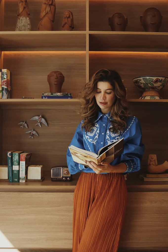 Thaisa Montenegro em ambiente decorado com peças artesanais, segurando um livro